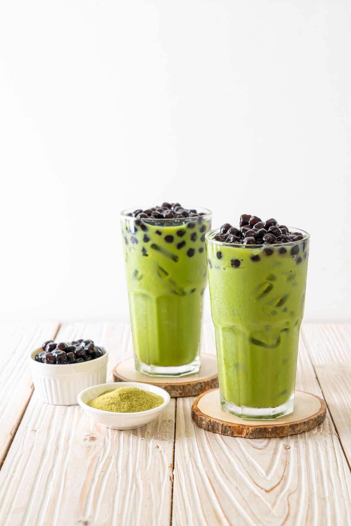 Two glasses of matcha boba tea with a side of tapioca pearls and matcha powder on a wooden surface against a white background.