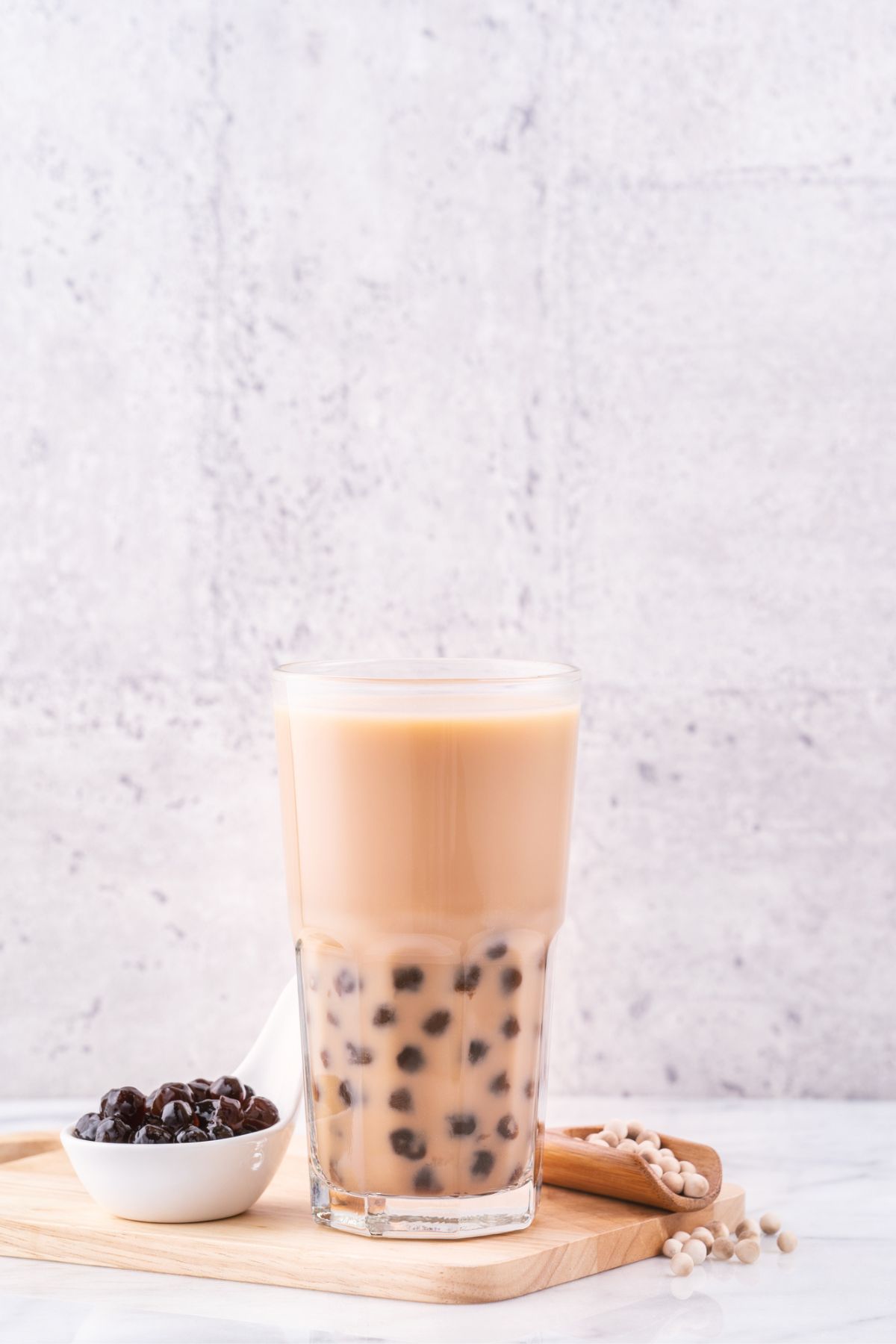 A tall glass of iced boba tea with tapioca pearls, accompanied by a small wooden plate of extra pearls and a bamboo scoop on a marble table against a textured wall.