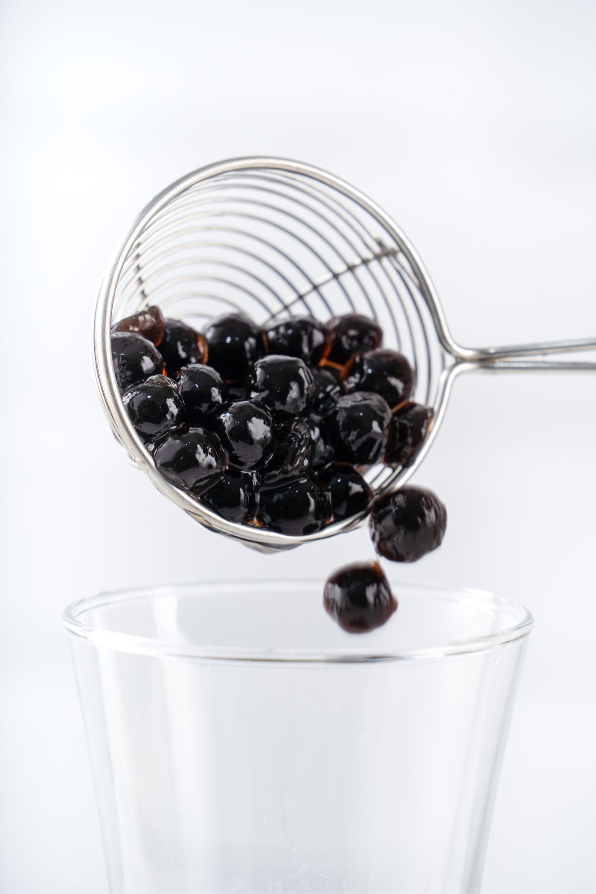 Black olives being drained from a slotted spoon over a clear glass bowl of boba tea.