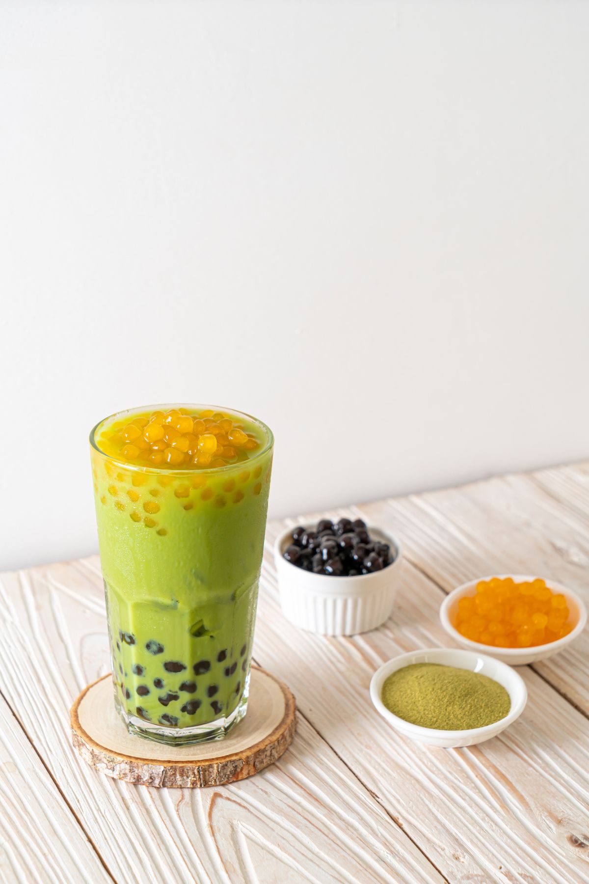 A tall glass of layered green matcha boba tea with tapioca pearls on a wooden coaster, accompanied by separate small dishes of black and golden tapioca pearls, and matcha powder on a light