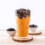 A glass of iced Yerba Mate bubble tea with black tapioca pearls on top, placed on a wooden coaster at home, accompanied by two small bowls of extra tapioca pearls on the side