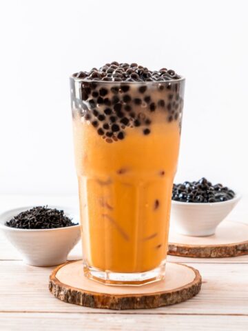 A glass of iced Yerba Mate bubble tea with black tapioca pearls on top, placed on a wooden coaster at home, accompanied by two small bowls of extra tapioca pearls on the side