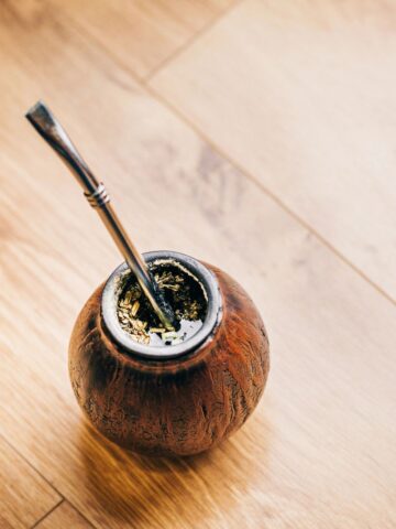 A traditional mate cup with a metal straw, known as a bombilla, filled with yerba mate for making at home, placed on a wooden floor.