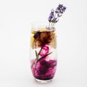 A glass of water infused with lemon slices, lavender, and a swirling dark purple simple syrup substitute, against a plain white background.